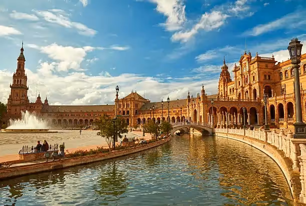 Elegant plaza with ornate, curved architecture, a central fountain, and a reflective canal under a blue sky. A few people stroll peacefully.