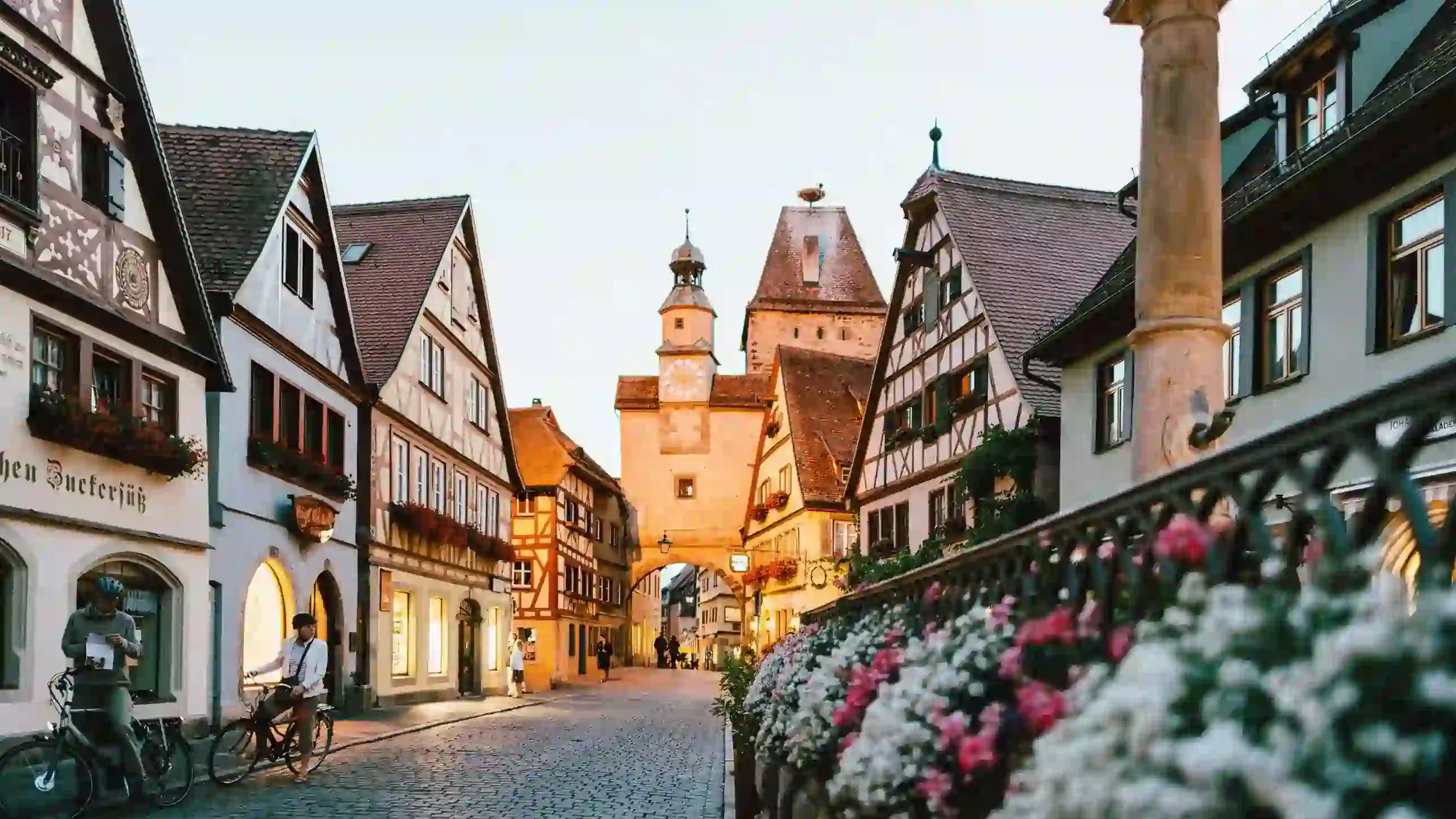 Quaint street in a medieval town with half-timbered houses, cobblestone path, and a distant clock tower. Floral railing adds charm and warmth.