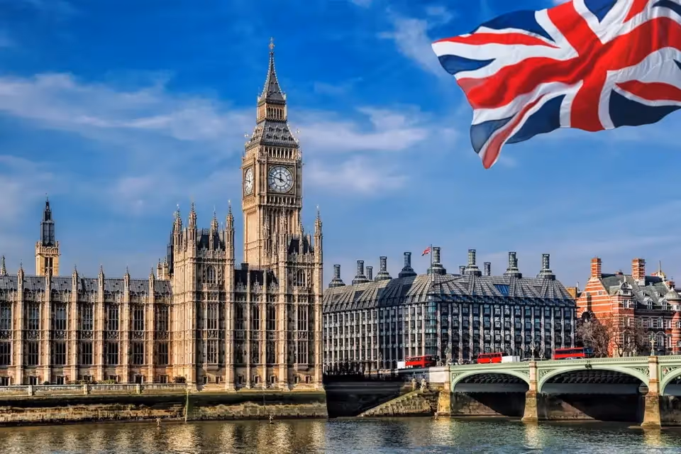 The image shows the iconic Big Ben and the Houses of Parliament in London, with a large Union Jack flag waving above. Red double-decker buses cross the bridge.