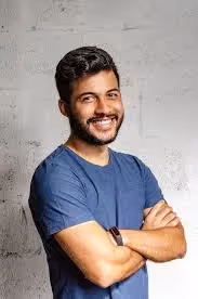 Un hombre sonriente, con el pelo corto y oscuro y barba, posa de pie frente a una pared de textura clara, vestido con una camiseta azul. Tiene los brazos cruzados con aire seguro.