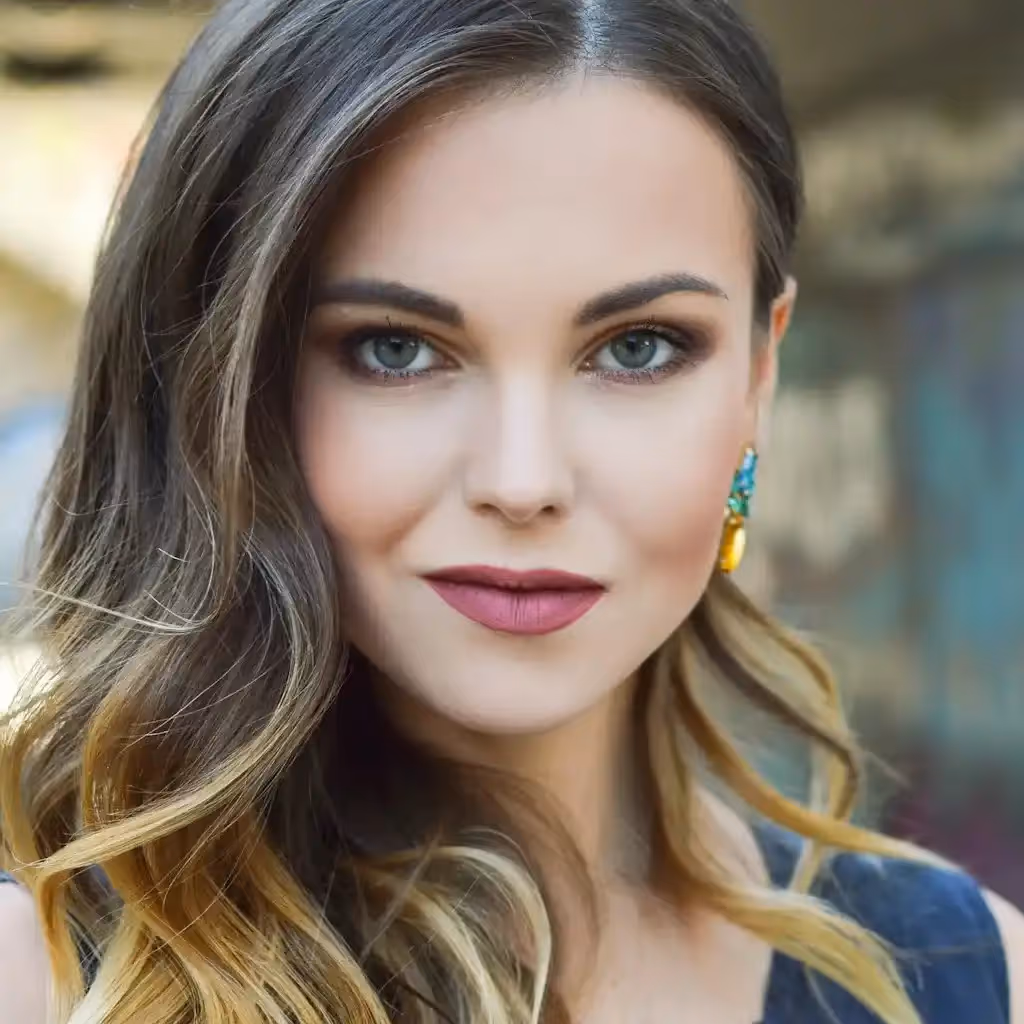 Une femme aux longs cheveux bruns et aux yeux bleus regarde l'objectif. Elle porte des boucles d'oreilles turquoise et un maquillage discret, rehaussé d'un rouge à lèvres rose, et dégage une grande assurance.