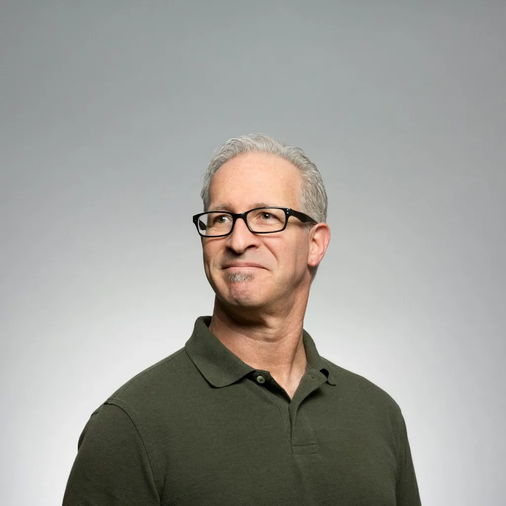 A man with gray hair and glasses in a dark green polo shirt looks slightly to the side; he appears thoughtful and confident, set against a plain gray background.