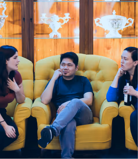 Three people sit on yellow armchairs in conversation. Their expressions are thoughtful. Ornate silver vases are displayed in the background.