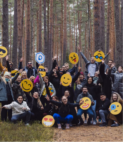 A joyful group of people in a forest, surrounded by tall trees, are enthusiastically posing with their arms raised. Emoji faces overlay their own, adding a playful and cheerful tone.
