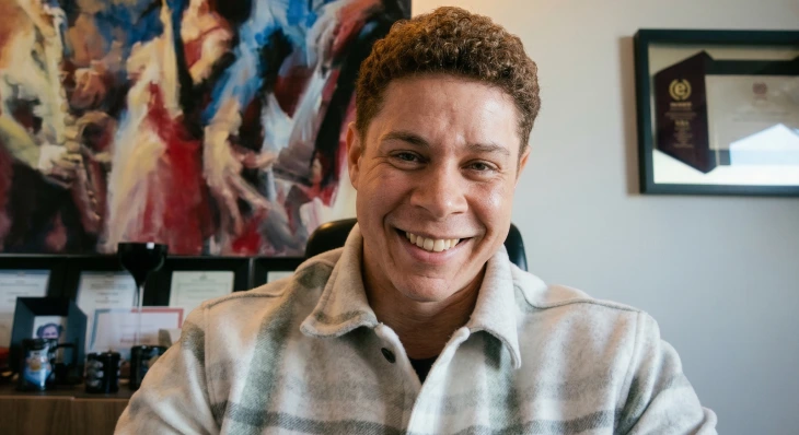 Smiling man with curly hair wearing a plaid shirt sitting in an office with framed art behind him.