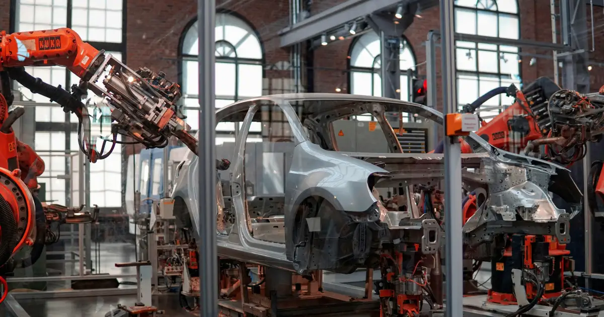 Robotic arms assembling a car frame inside a factory with large windows and industrial equipment.