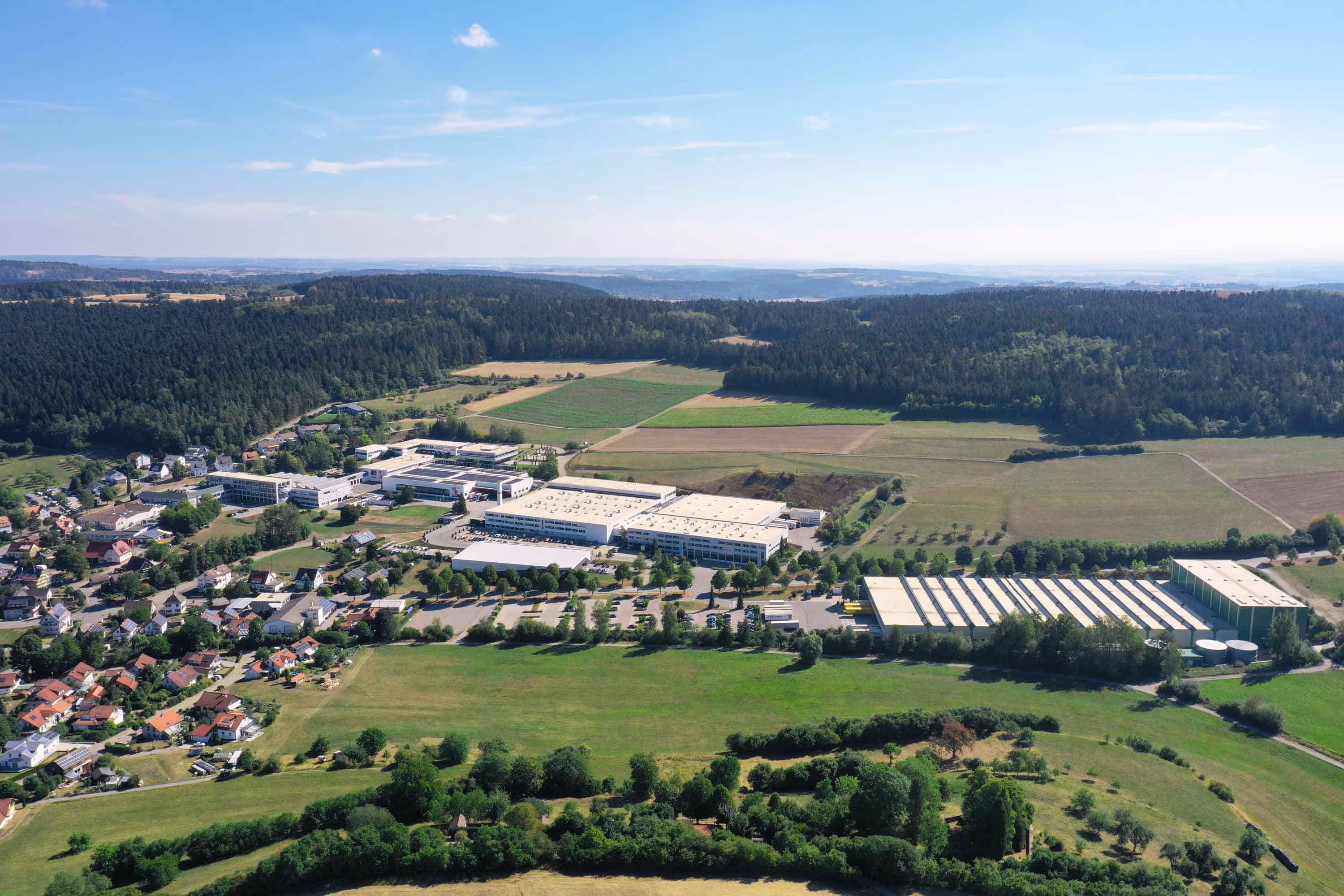 Aerial view of a manufacturing campus with large buildings surrounded by fields, forests, and a small residential area.