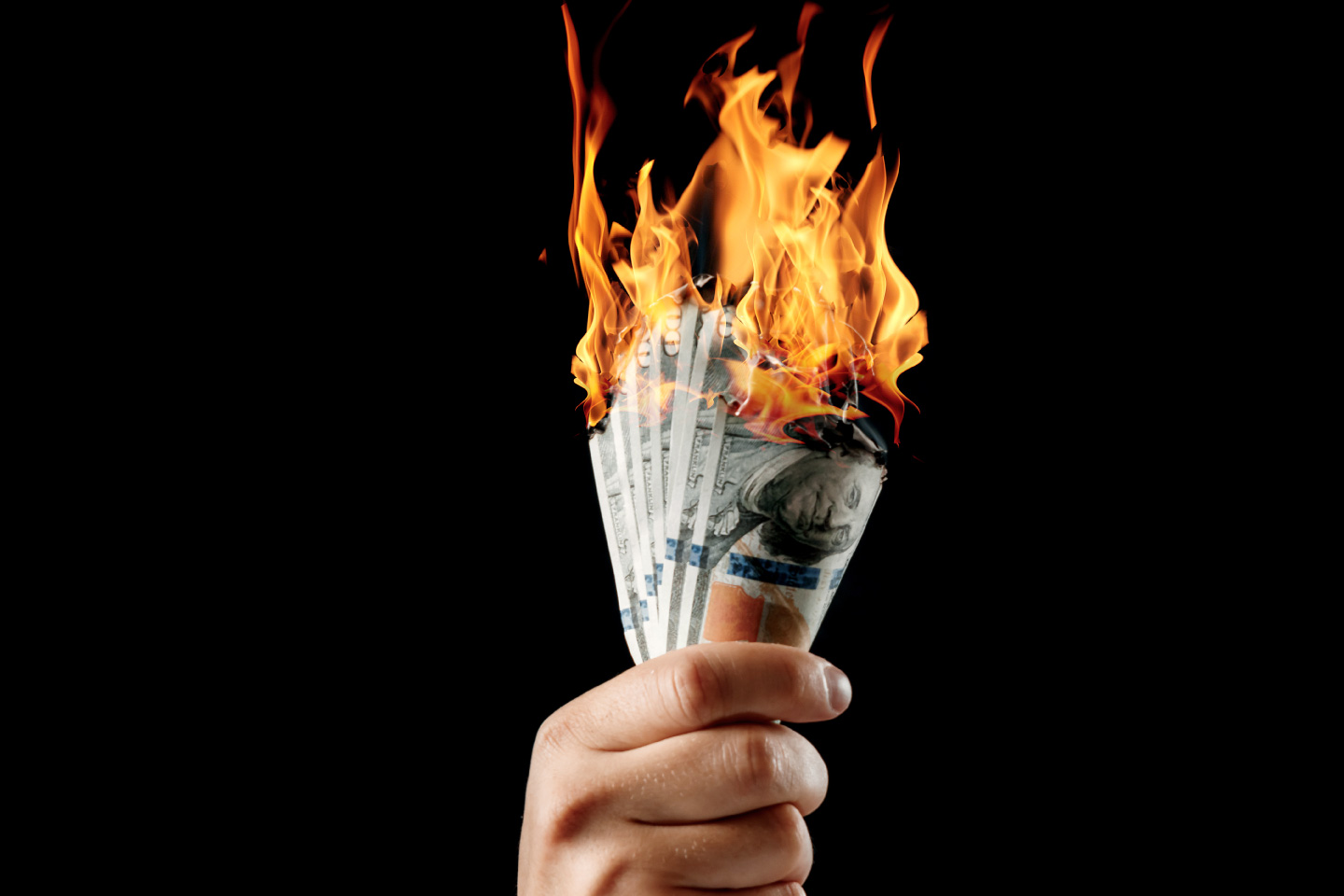 A hand holding a stack of seven burning one hundred dollar bills