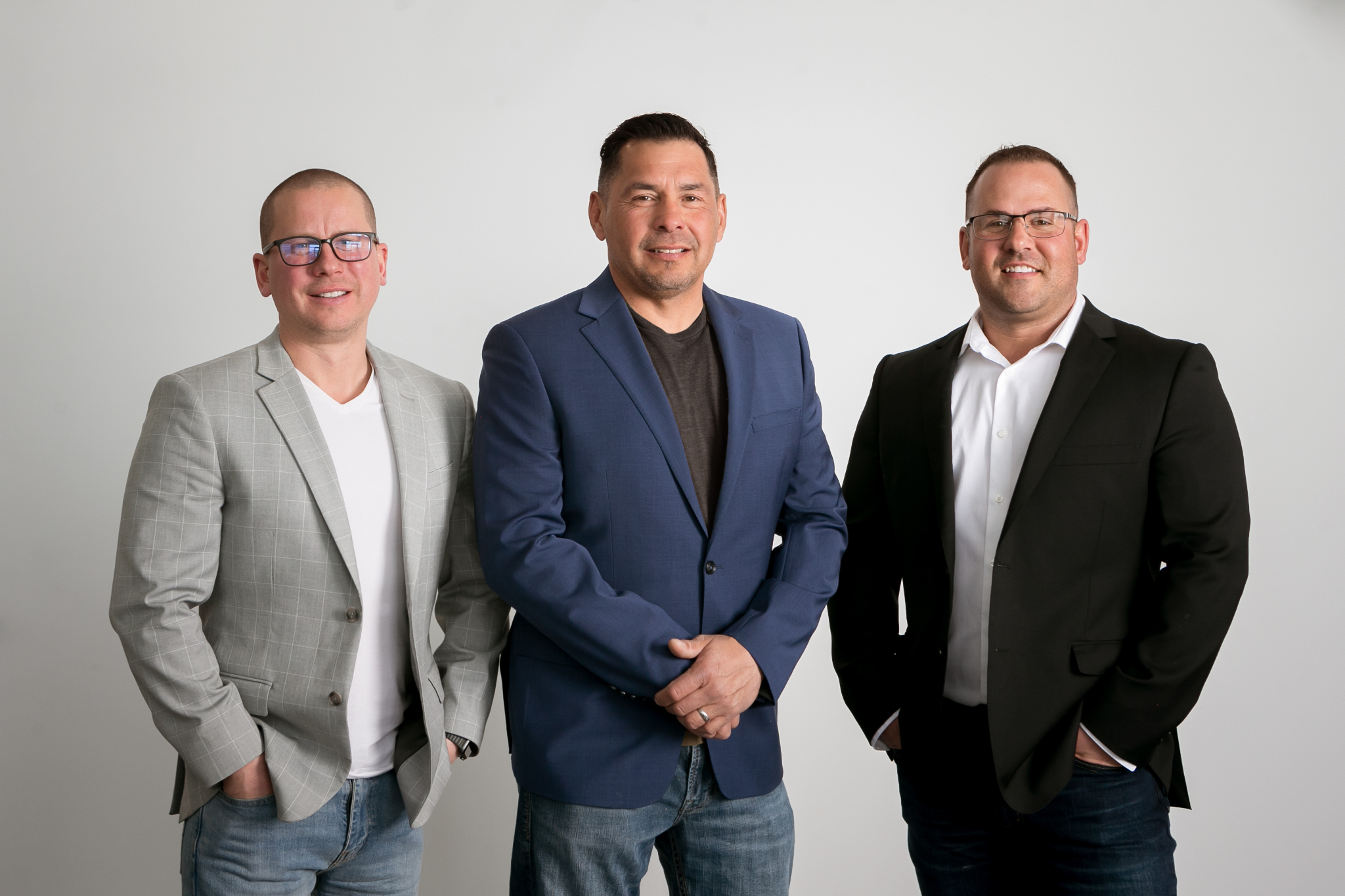 Three men standing against a plain light gray background wearing casual business attire, including blazers and jeans, all with their hands either in pockets or folded in front.