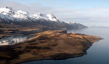 Mountainous landscape with snow-capped peaks reflecting on a calm lake beside a grassy peninsula.
