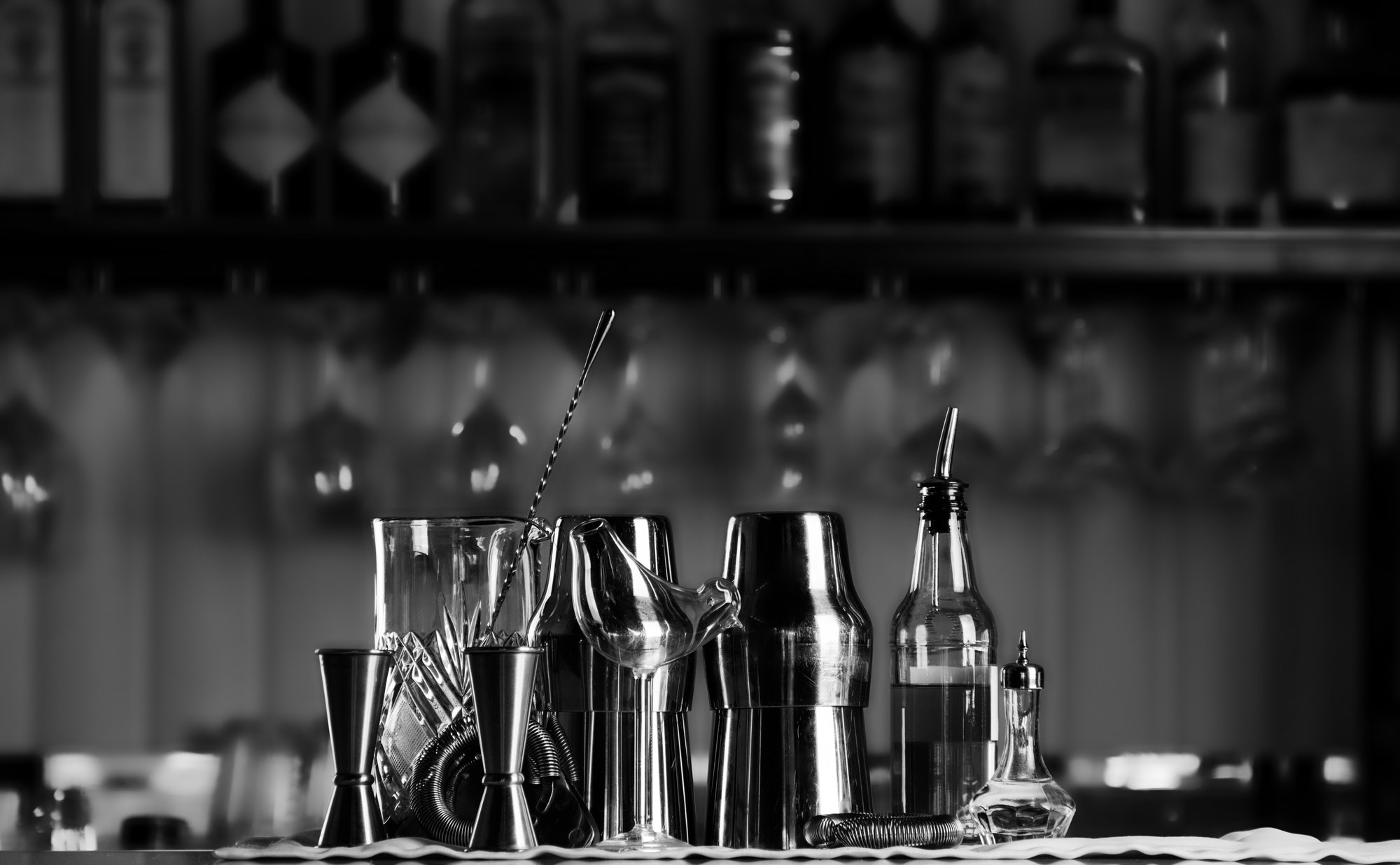 Bartender's tools — jiggers, a mixing glass, bar spoon, shakers, a strainer and a bitters bottle — lined up on a bar counter in front of a backlit bottle shelf.