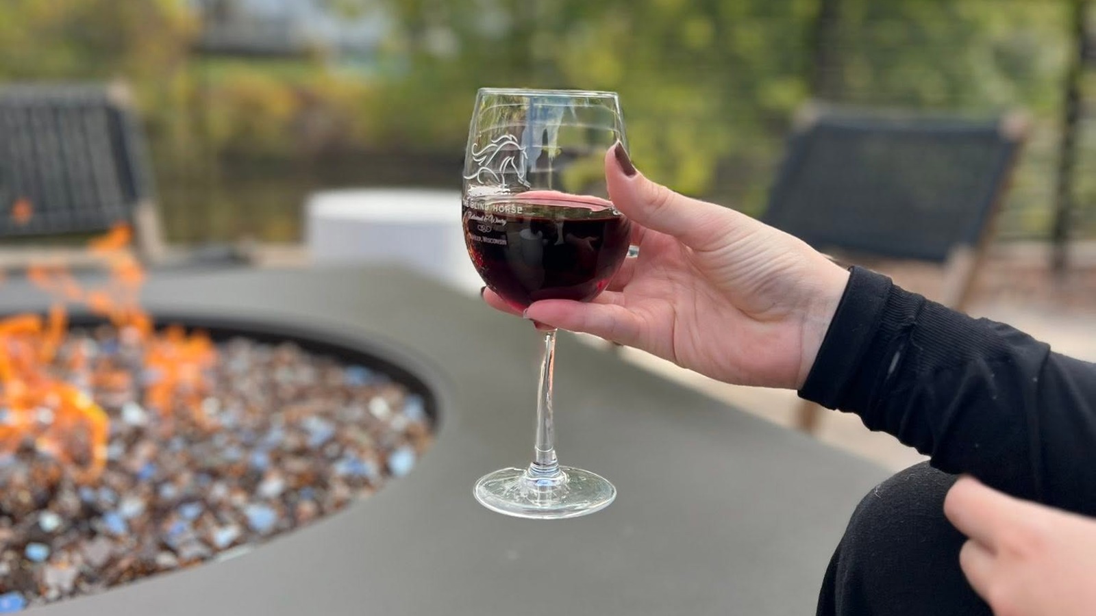 A glass of red wine etched with The Blind Horse logo, held beside a fire pit on the patio.