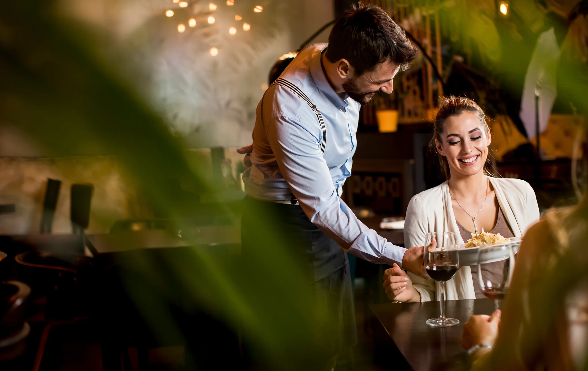 Un mesero sirve un platillo a una comensal sonriente en la mesa de un restaurante durante el servicio de la noche