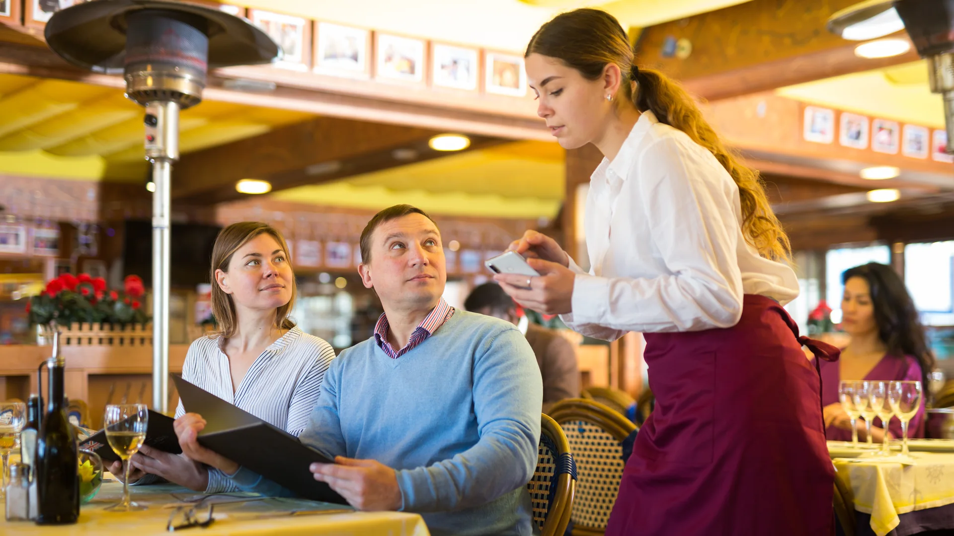 A server takes an order at the table on a phone while two seated guests look up from the menu.
