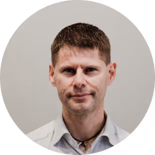 Headshot of a man with short brown hair wearing a light-colored collared shirt and a necklace with a dark cord against a neutral background.