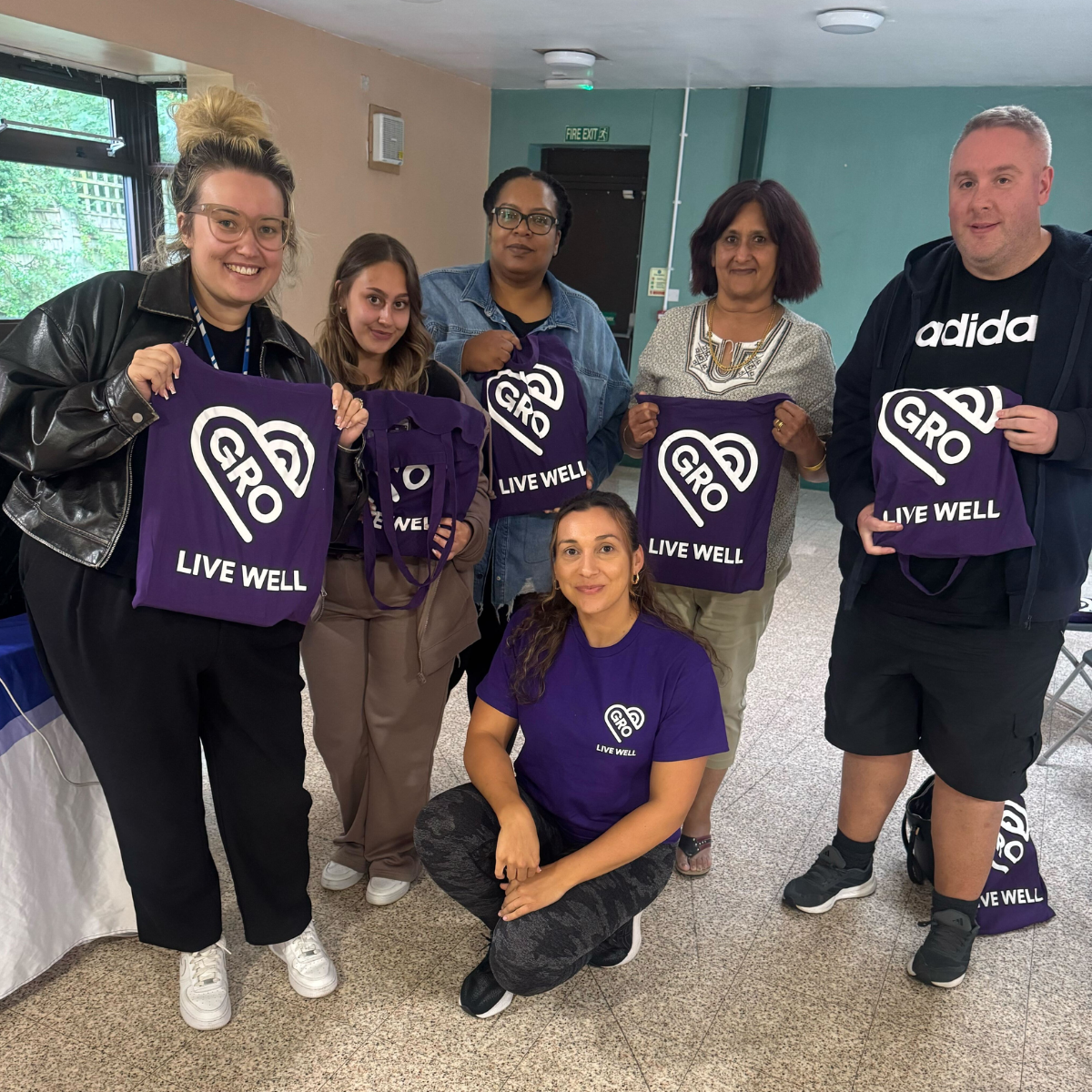 Group of five people smiling and holding purple GRO Live Well tote bags, with one woman kneeling wearing a matching purple GRO Live Well T-shirt.