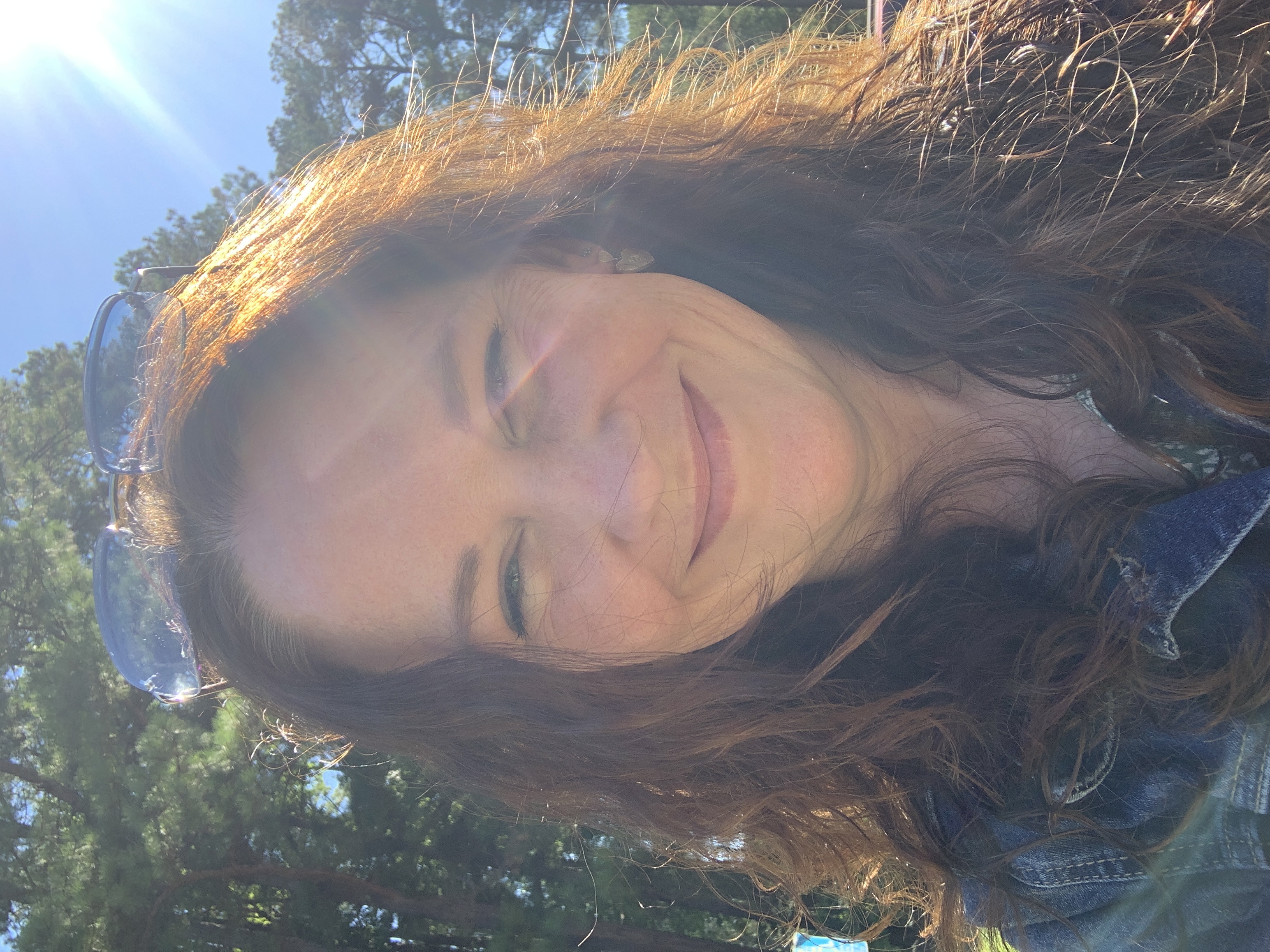 Smiling woman with long curly hair wearing a denim jacket and sunglasses on her head outdoors with sunlight filtering through trees.