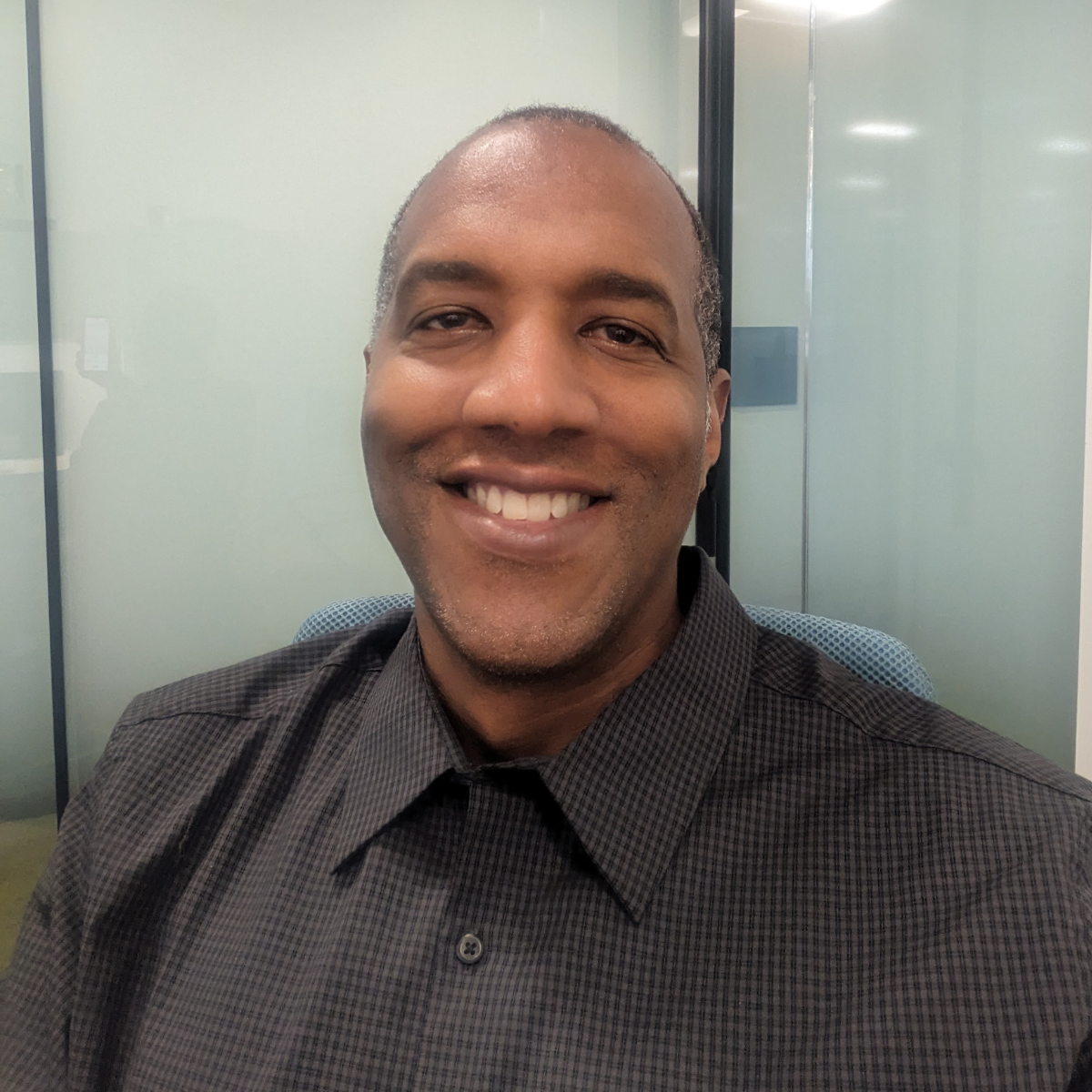 Smiling middle-aged man wearing a dark checkered shirt sitting in an office chair.