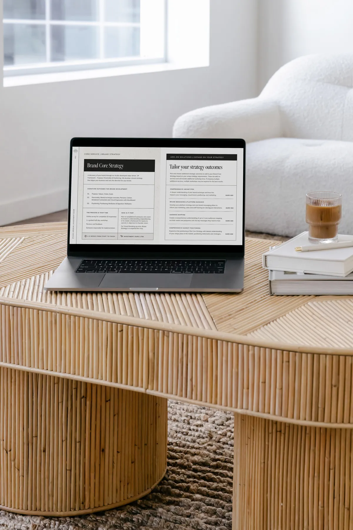 Laptop on a bamboo coffee table displaying a webpage titled 'How we can work together' outlining brand strategy, visual branding, website creation, and brand activation services; next to a glass of coffee and stacked books.