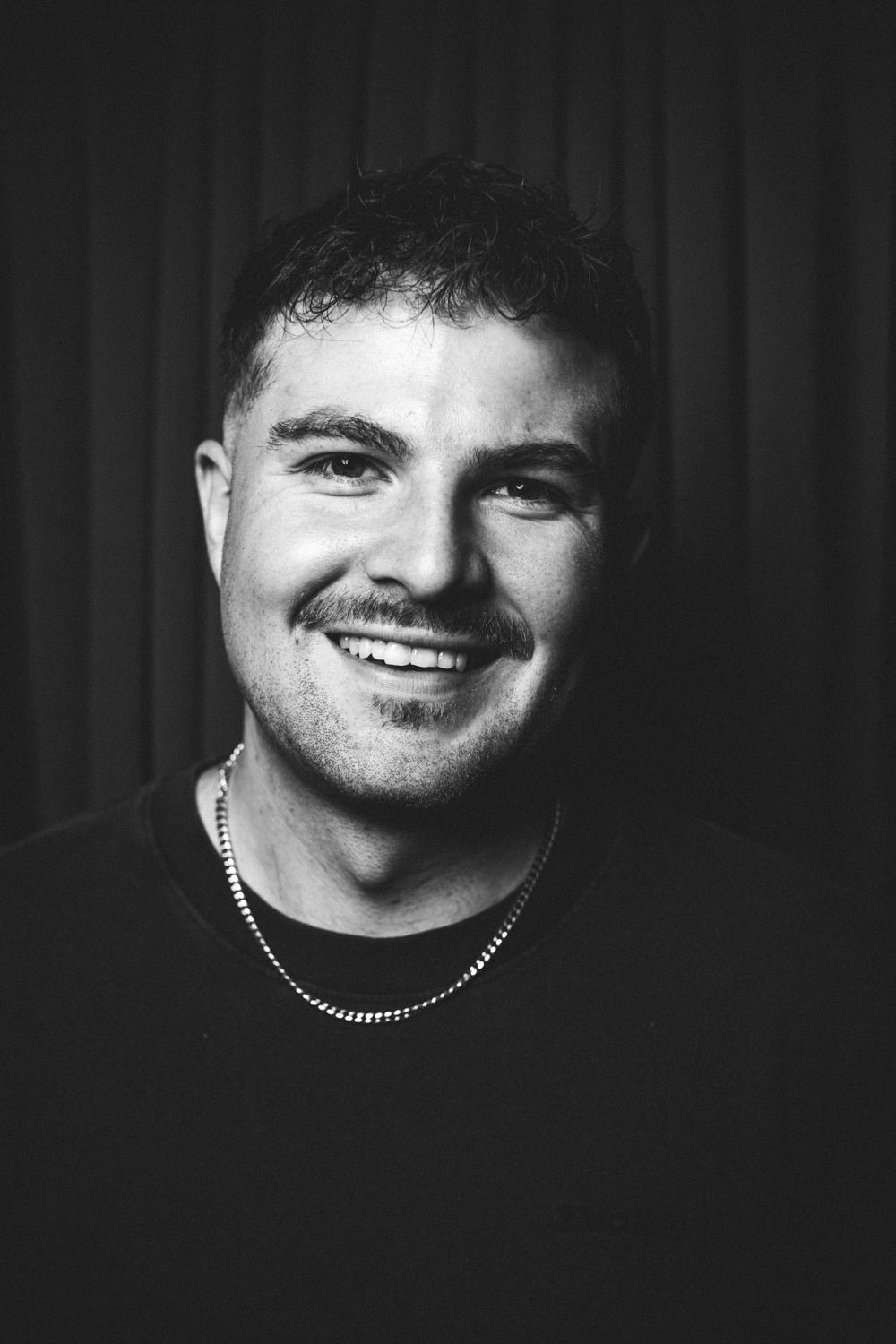 Black and white portrait of a smiling man with short curly hair, a mustache, and a chain necklace.