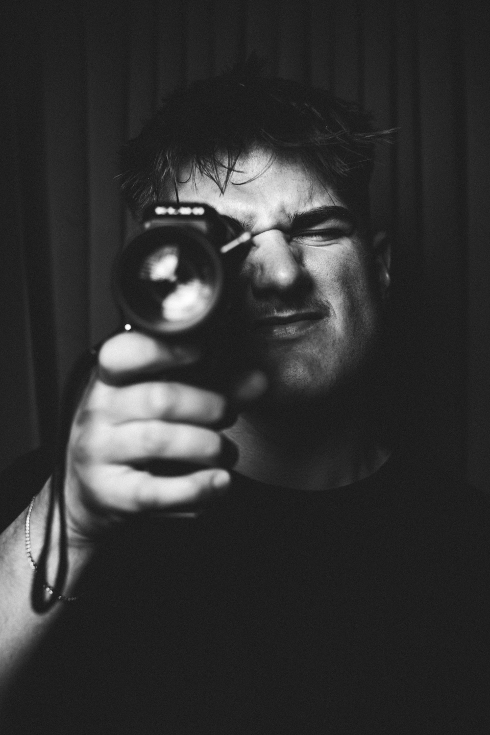 Black and white photo of a man aiming a camera directly at the viewer, one eye closed.