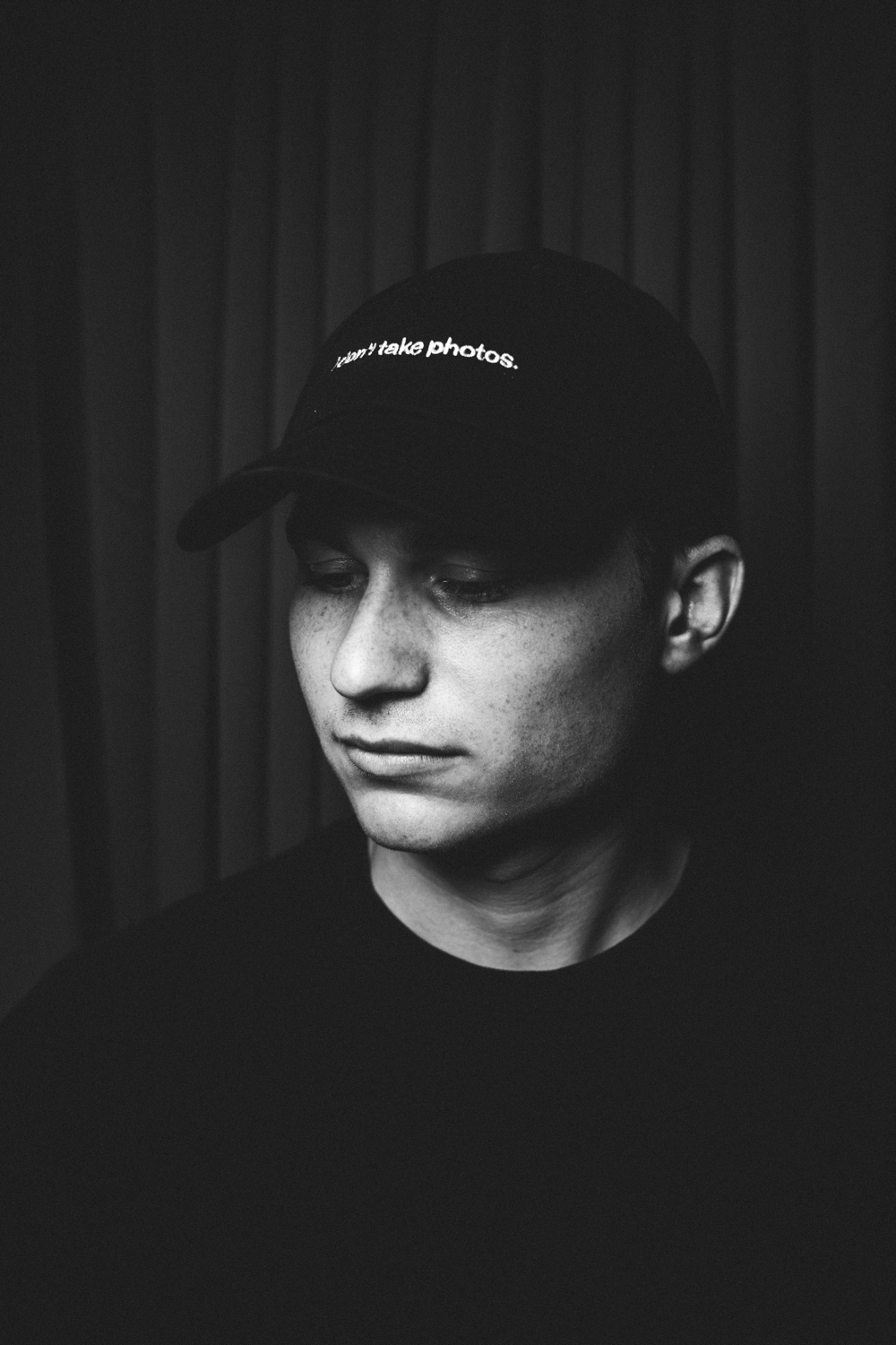 Black and white portrait of a young man wearing a cap that says 'I don't take photos.' looking down.