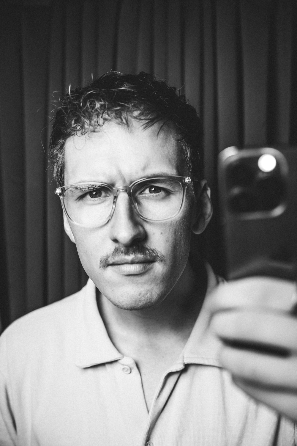 Man with curly hair, mustache, and clear glasses taking a black-and-white selfie in front of a curtain.