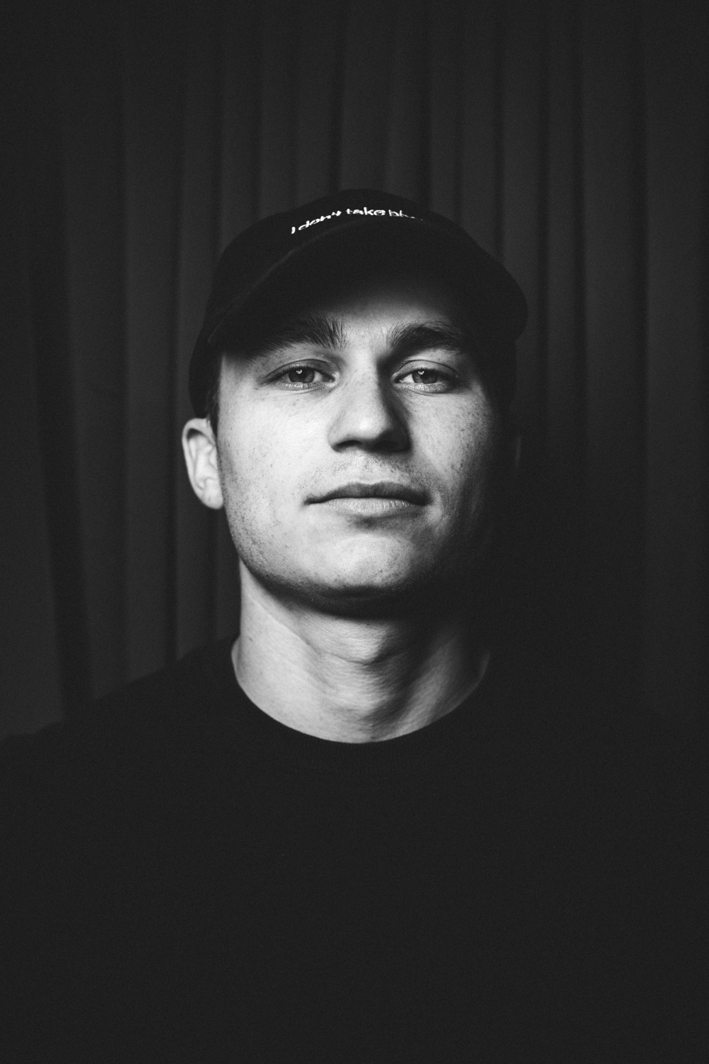 Black-and-white portrait of a young man wearing a cap with a neutral expression and a dark background.