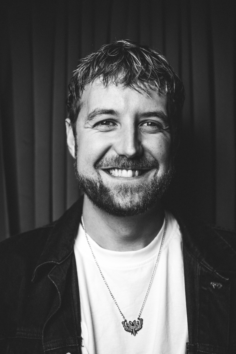 Smiling man with short beard and necklace, wearing a denim jacket and white shirt, black curtain backdrop.