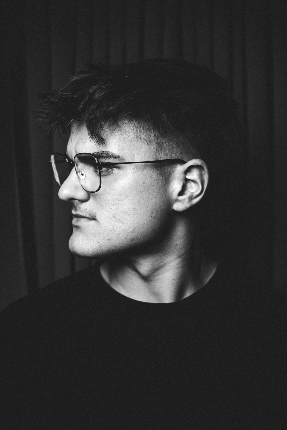 Black and white portrait of a young man with glasses looking to his left against a dark background.