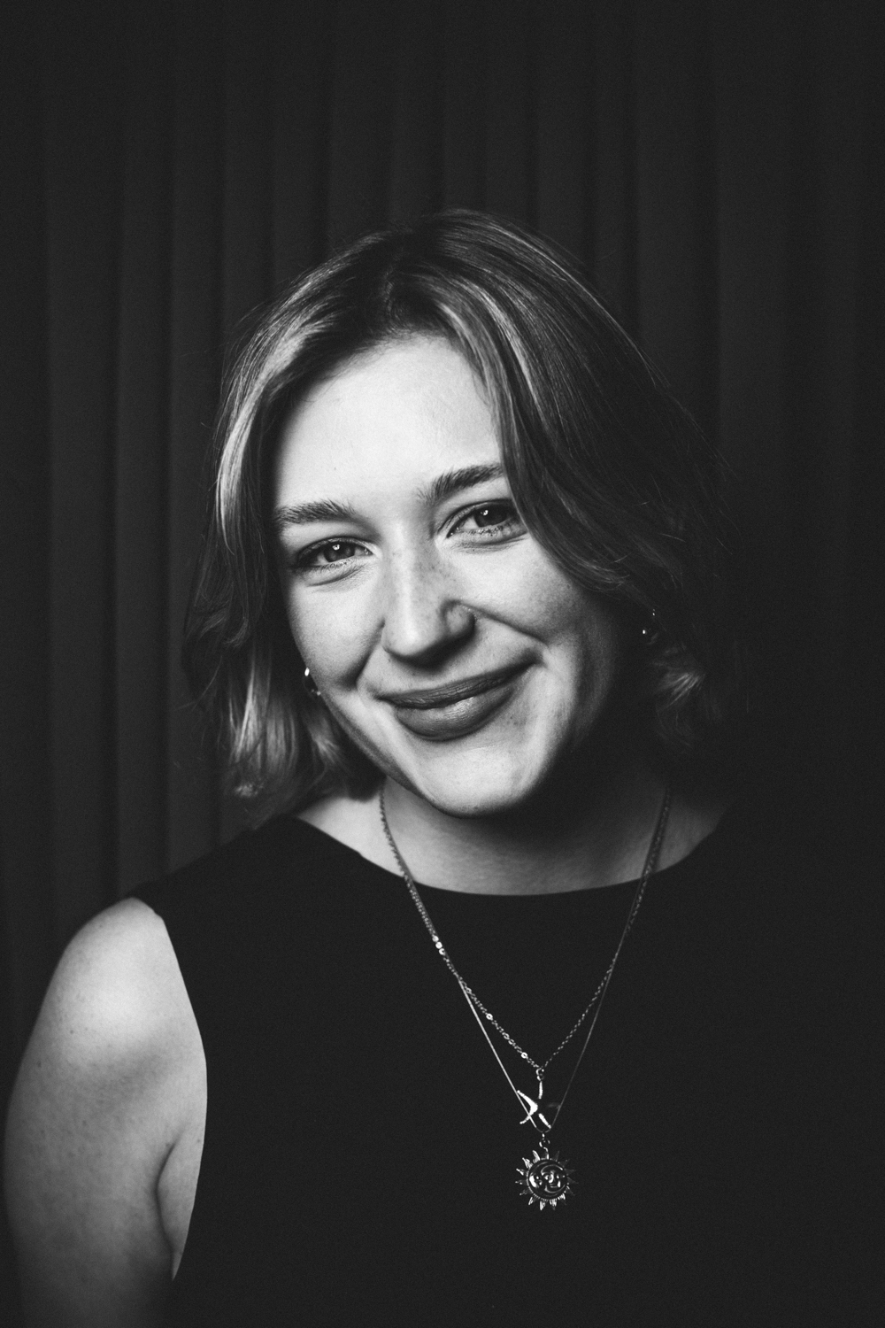 Black and white portrait of a smiling woman with short hair wearing a sun pendant necklace.