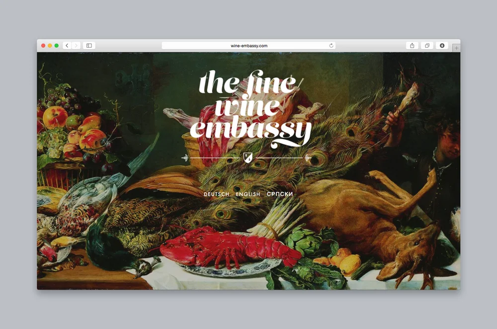 Ein Screenshot eines Desktop-Webbrowsers, der die Landingpage der Website „The Fine Wine Embassy“ zeigt. Der Hintergrund zeigt ein klassisches, detailreiches Stillleben im Barockstil, das ein üppiges Festmahl mit einem Hirsch, einem Hummer auf einem Teller, einem Pfau, verschiedenen Früchten wie Weintrauben und Äpfeln sowie anderem Wild darstellt. Eine große, stilisierte weiße Serifenschrift in der Mitte lautet „the fine wine embassy“, darunter sind die Sprachoptionen für Deutsch, Englisch und Serbisch aufgeführt.
