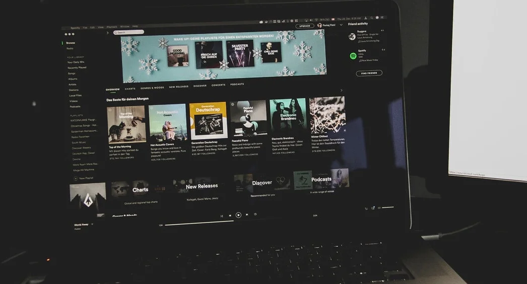 Eine Nahaufnahme eines Laptop-Bildschirms, der die Benutzeroberfläche der Spotify-Desktop-App im Dunkelmodus anzeigt. Der Bildschirm zeigt verschiedene Musik-Playlist-Karten, ein winterliches Banner am oberen Rand und ein Seitenleisten-Navigationsmenü. Rechts ist ein Teil eines externen Monitors zu sehen, und die Laptop-Tastatur befindet sich im Vordergrund bei gedimmtem Licht.