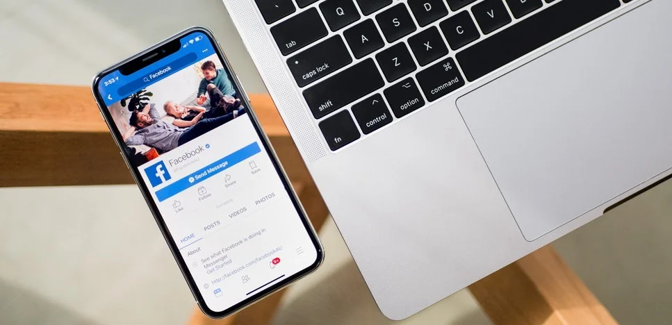 Eine Draufsicht auf ein iPhone, das die Profilseite der Facebook-Mobil-App anzeigt, neben einem geöffneten silbernen Laptop auf einer Holzoberfläche.