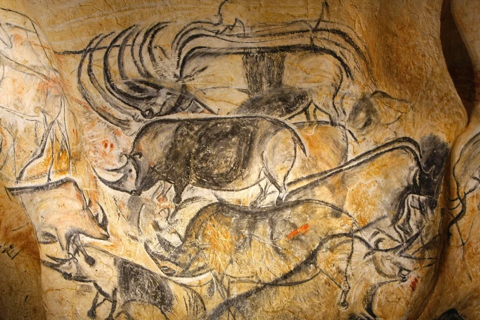 Eine prähistorische Höhlenmalerei, die eine Herde von Wollnashörnern und ein Mammut darstellt, gezeichnet mit schwarzer Holzkohle auf einer unebenen, ochserfarbenen Steinwand. Die Tiere sind mit dynamischen Umrissen und schattierten Körpern skizziert, was ein Gefühl von Bewegung vermittelt.