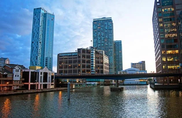 Une vue sur le front de mer avec des gratte-ciels et des bâtiments modernes, dont l'hôtel Britannia International, surplombant un bassin calme à Canary Wharf, Londres, sous un ciel de crépuscule avec un pont traversant l'eau.