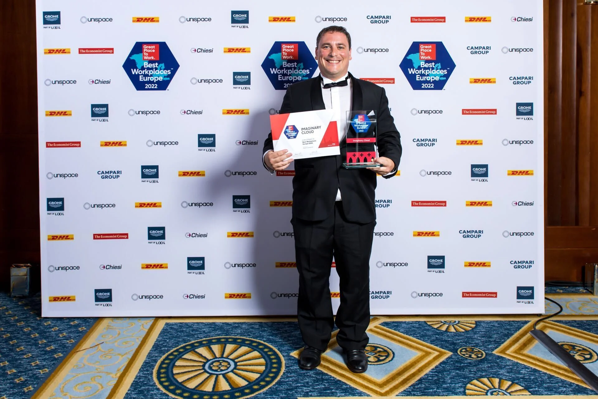 Un homme souriant en smoking noir et nœud papillon se tient devant un photocall blanc couvert de logos répétitifs tels que DHL, Unispace et Grohe. Il tient d'une main un certificat « Best Workplaces Europe 2022 » décerné à « Imaginary Cloud » et de l'autre un trophée en verre transparent assorti.