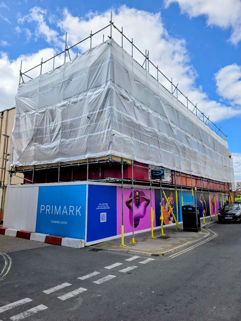 Building under construction wrapped in white scaffolding with a blue Primark 'Coming Soon' advertisement along the lower perimeter.