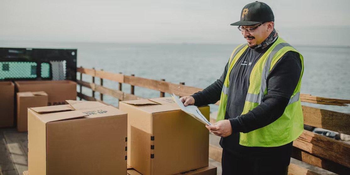 A Curri delivery driver wearing a high-visibility vest checks paperwork while standing on a truck bed surrounded by large boxes, with a body of water in the background.