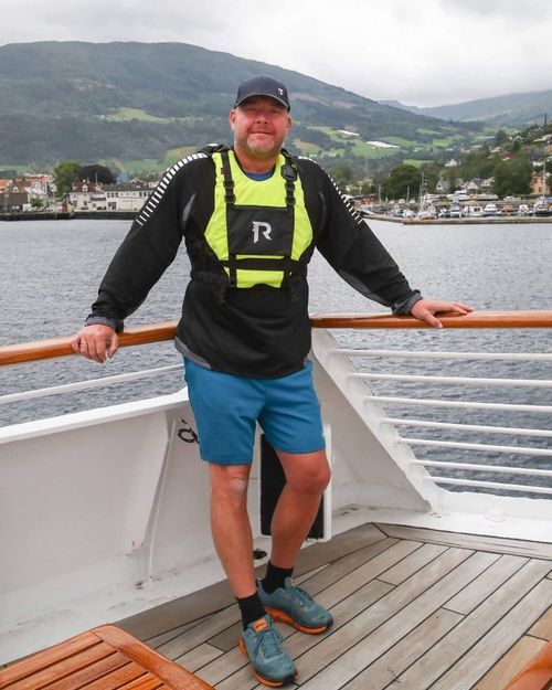 Man in a black jacket and blue shorts standing on a wooden deck of a boat with mountains and a marina in the background.