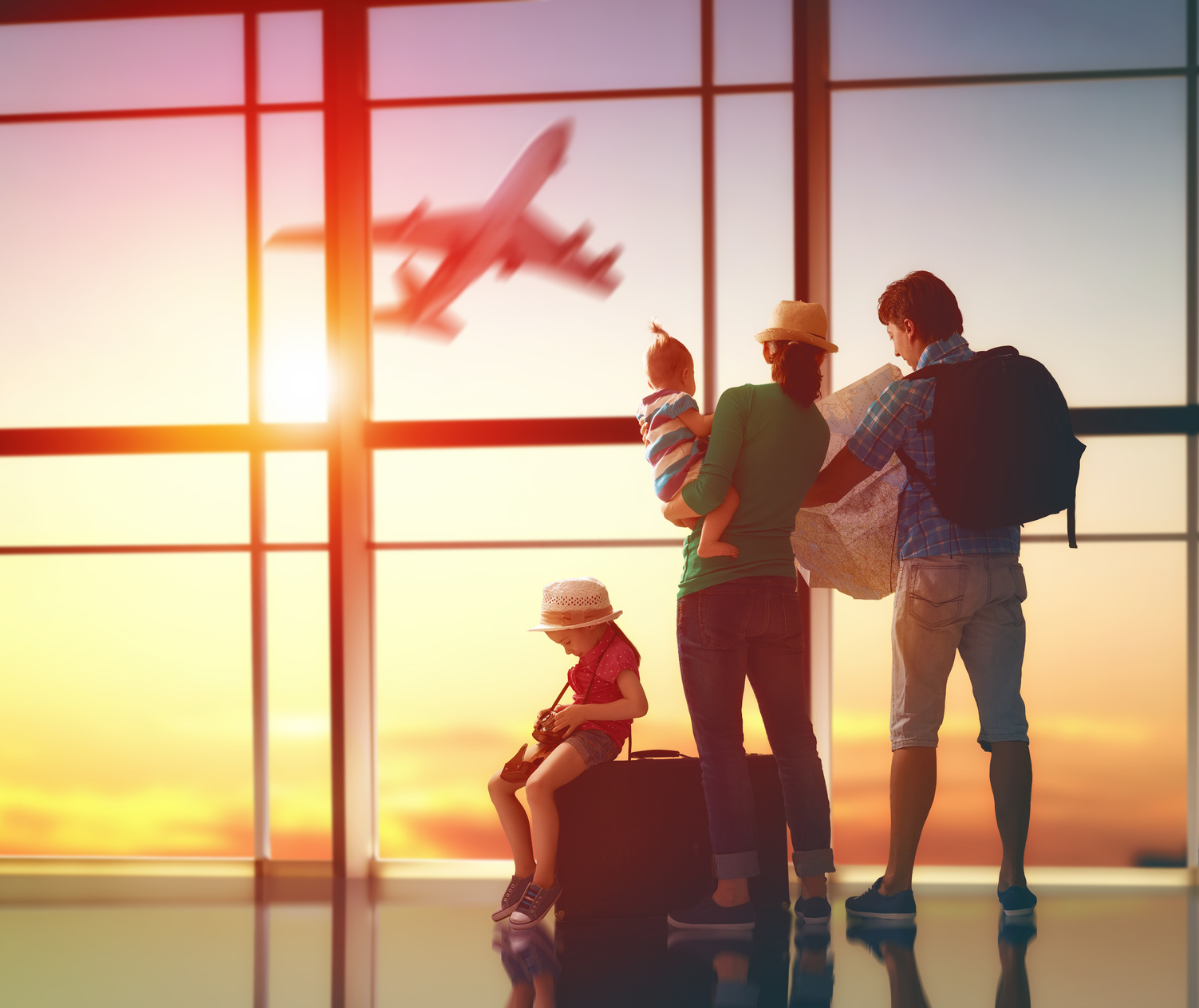 Family of four at an airport terminal at sunset, watching a plane take off outside the window.