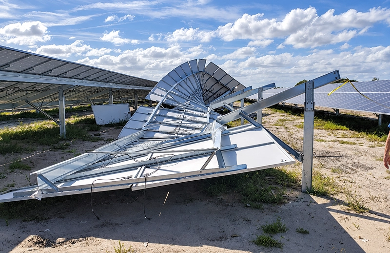 A damaged solar panel array lies bent and collapsed in a sunlit solar farm, with intact panels behind it under a bright, cloud-filled sky.