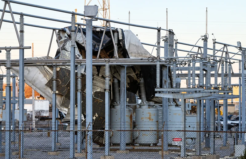 Damaged electrical substation equipment stands behind a chain-link fence, with torn metal panels and tangled cables conveying a stark, industrial sense of destruction.