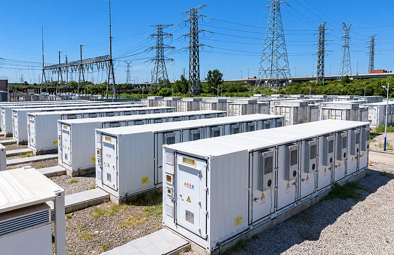Rows of white electrical switchgear cabinets fill an outdoor substation beneath towering power lines, conveying a bright, orderly industrial scene.