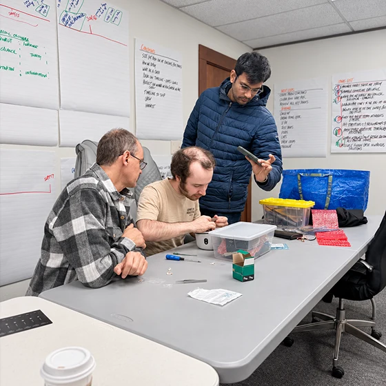 Three men collaborate on a tabletop project in a classroom, examining a small device and materials beneath walls covered with notes; focused, hands-on teamwork.