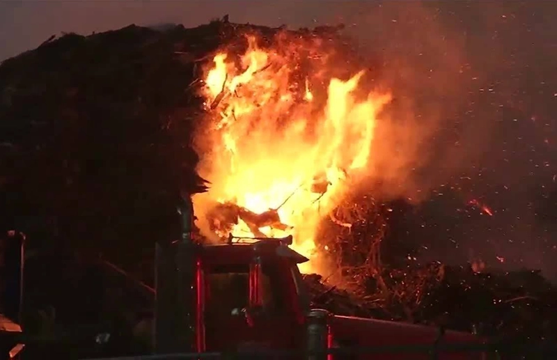 A massive nighttime fire engulfs a tree-covered hillside, sending bright orange flames and sparks into the smoky sky above a dark vehicle. The scene feels dangerous and urgent.