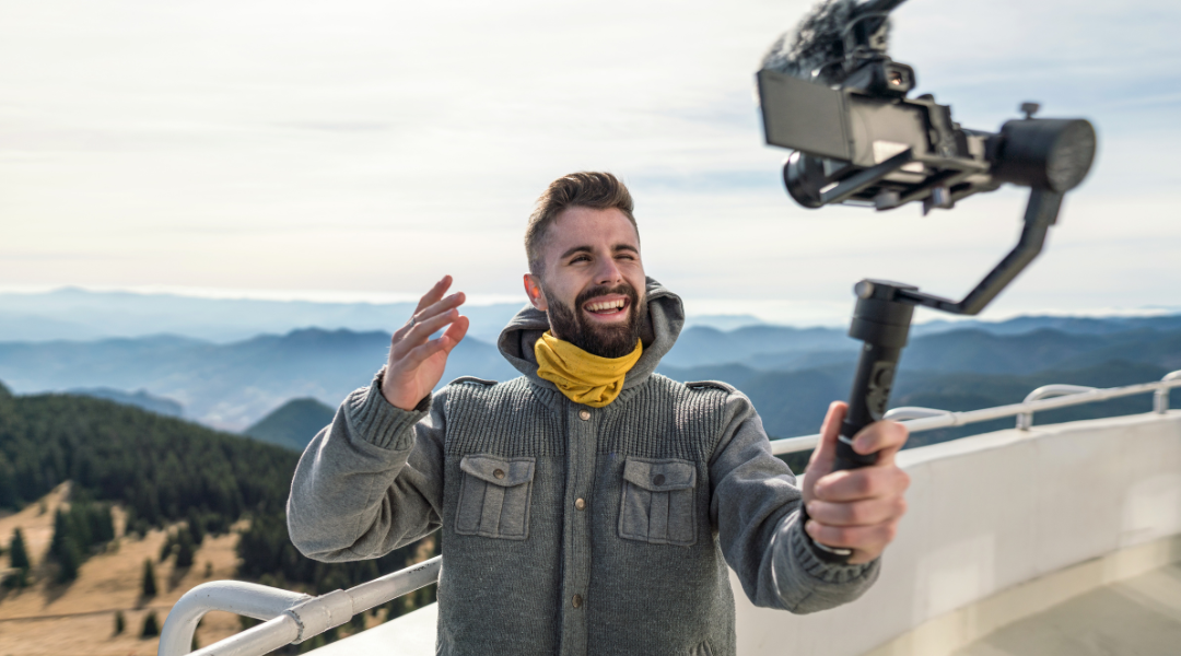 A man holding a camera filming himself on top of a balcony overlooking mountains and scenery