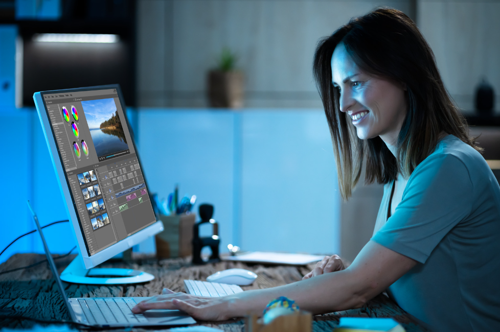 A woman is sitting in front of the desk, looking at the screen on her laptop. On the side, she has another monitor. It shows the editing screen. She is smiling.