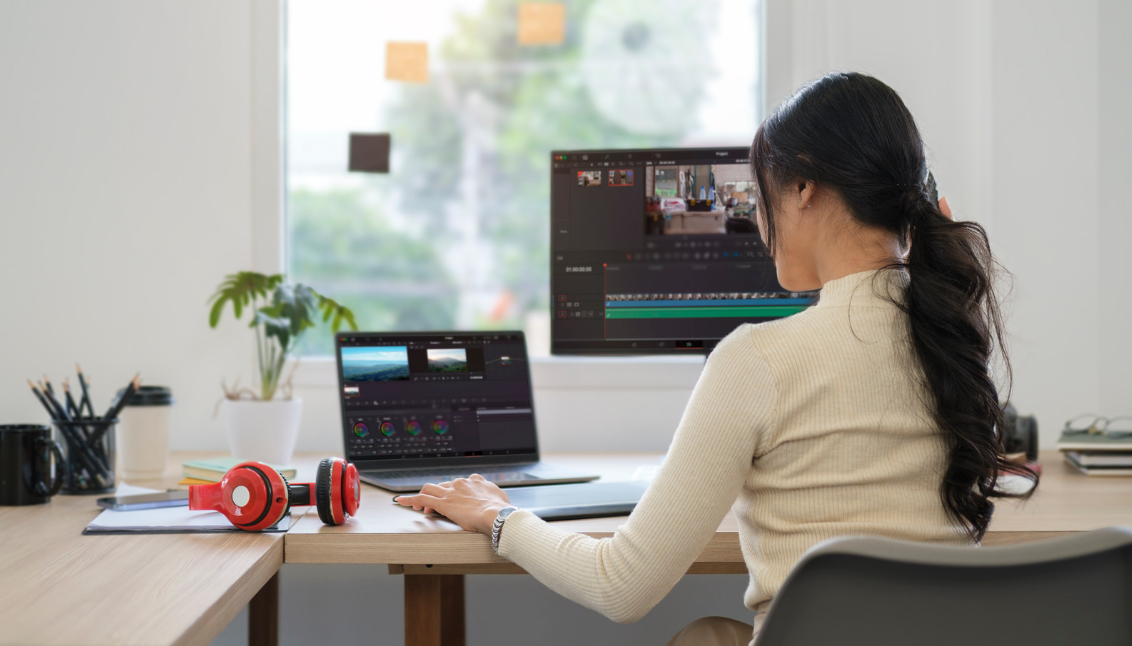 A woman sitting in front of the desk editing videos on her laptop. It has another monitor on the side.