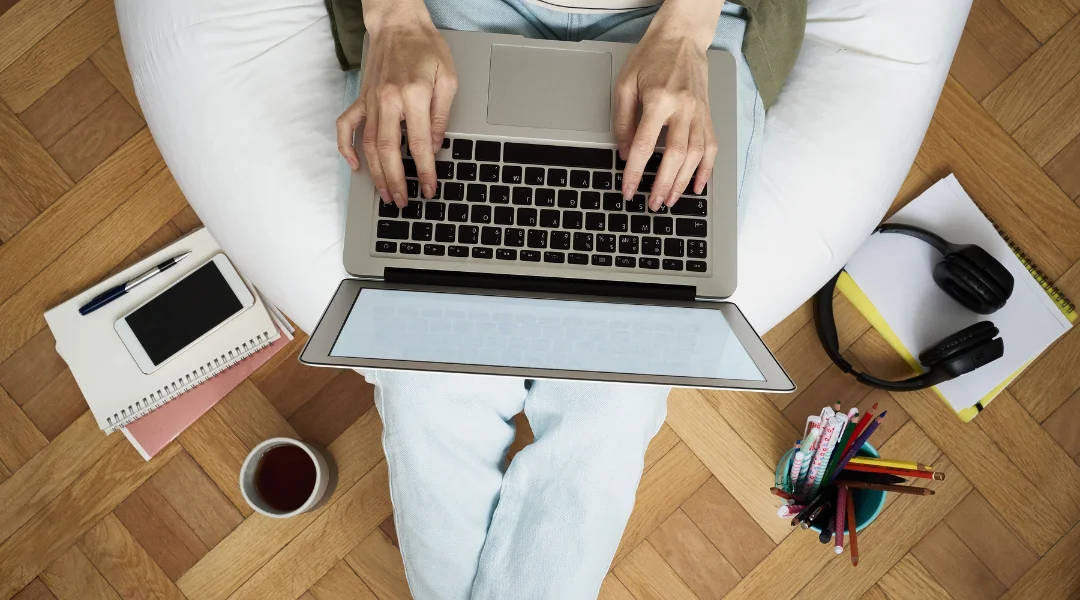 A woman sitting on a bean chair with her PC, coffee, headphones, notebooks, and colored pens